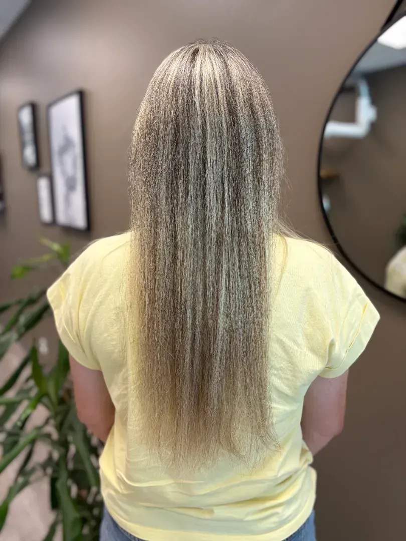 Person with long, straight, light brown hair, wearing a yellow shirt, stands in a salon.