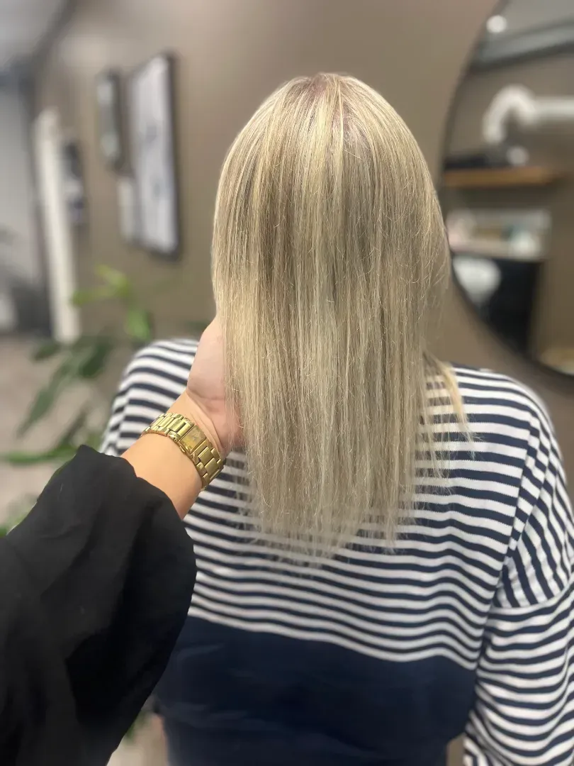 Woman with blonde, crimped hair at a salon. Someone's hand touches her hair. She wears a striped top.