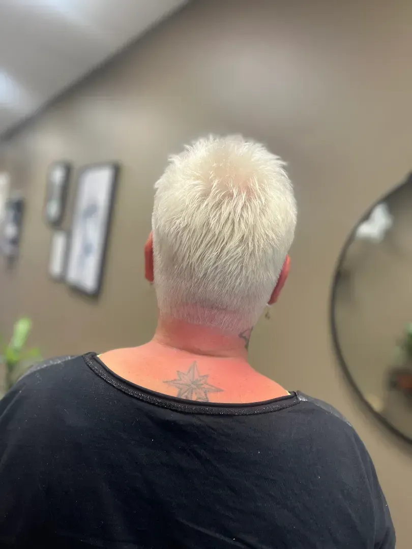 Back of a person with short, platinum blonde hair in a salon setting. They are wearing a black top and have a neck tattoo.
