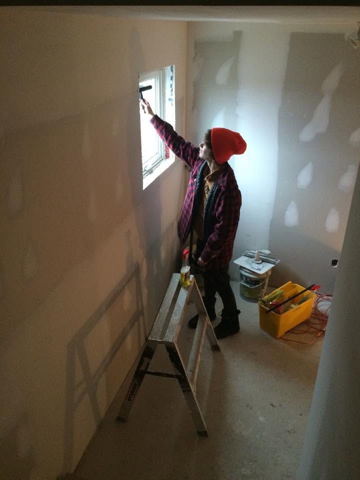 Person on a ladder painting a wall around a small window in an empty room
