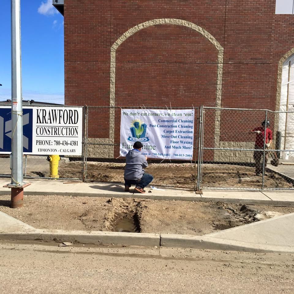 Child in a gray coat crouches by a chain-link fence with signs outside a brick building.