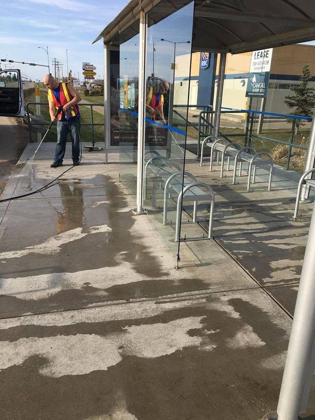 Worker power-washing a wet sidewalk beside a bus stop shelter and metal benches
