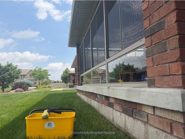 Yellow trash bin on grass beside a brick building with large windows on a sunny day