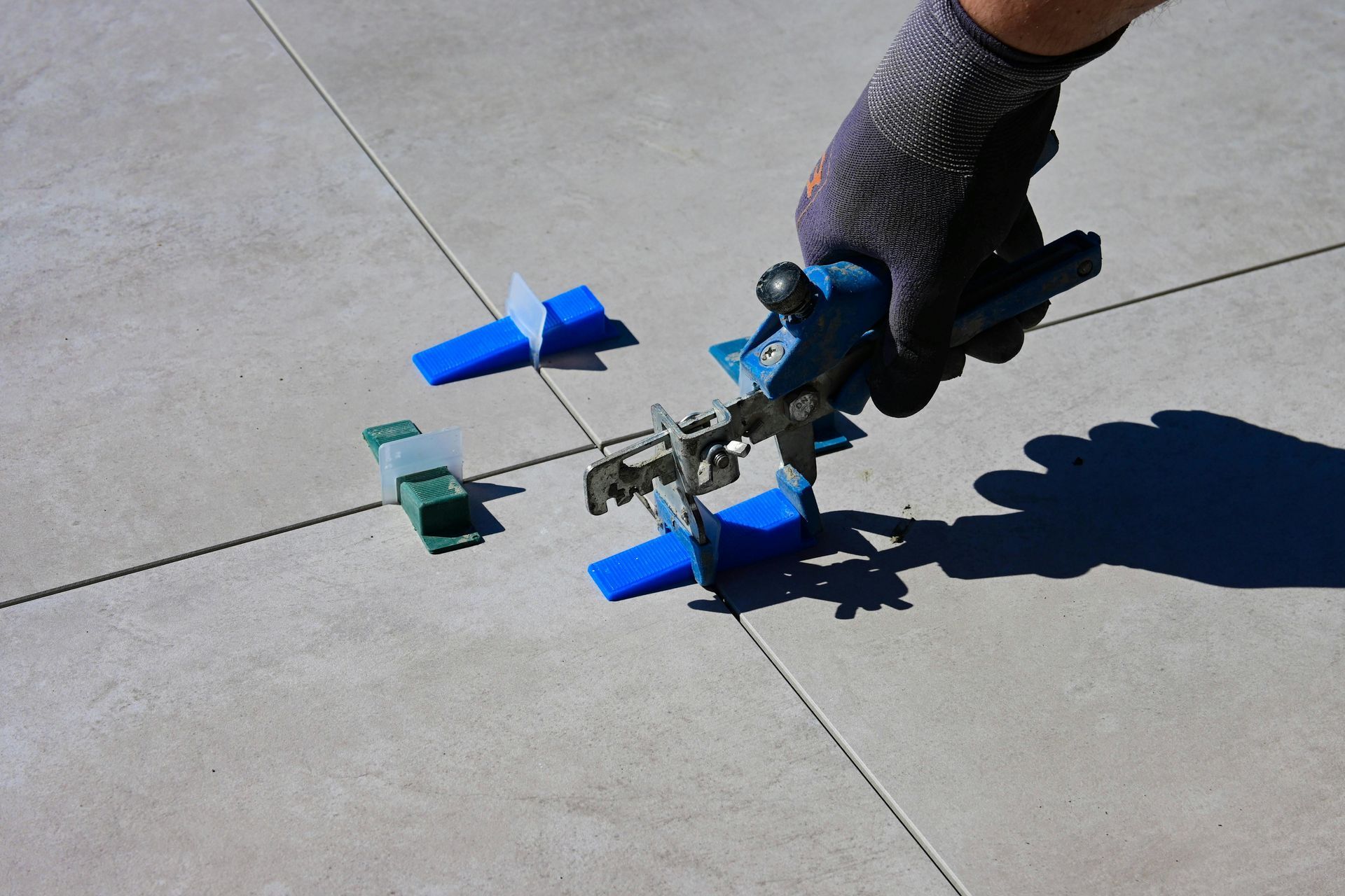 Hand using a tile leveling system on large gray floor tiles with blue and green plastic pieces.