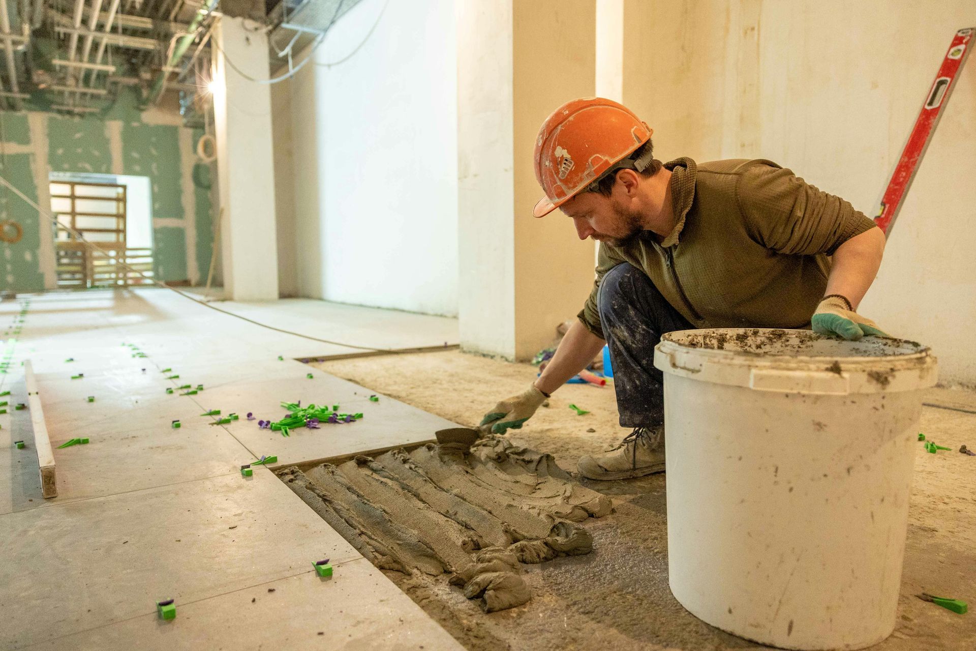 Man in hard hat tiling a floor with a trowel, bucket of mortar nearby; construction site.