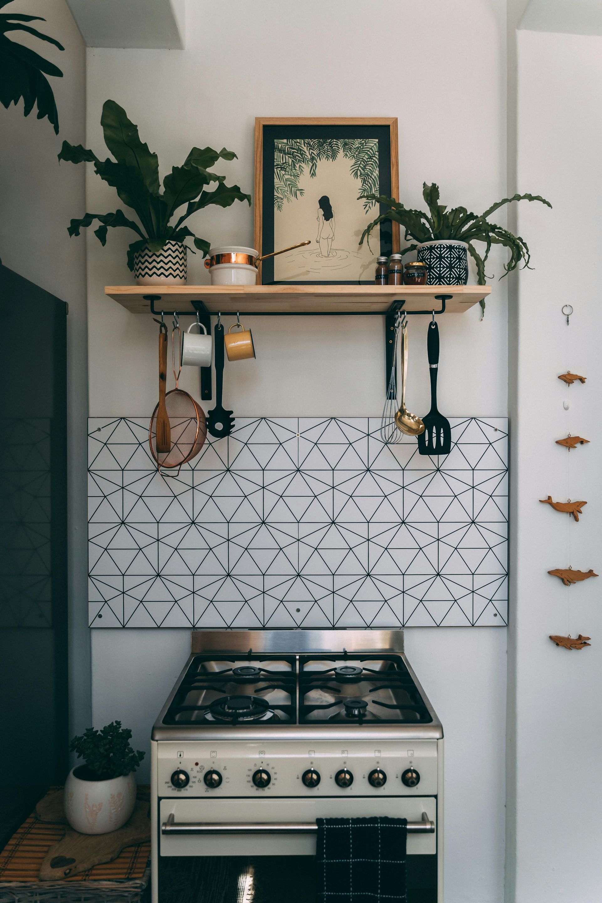 Kitchen with stove, tiled backsplash, shelf with plants and artwork, utensils hanging below.