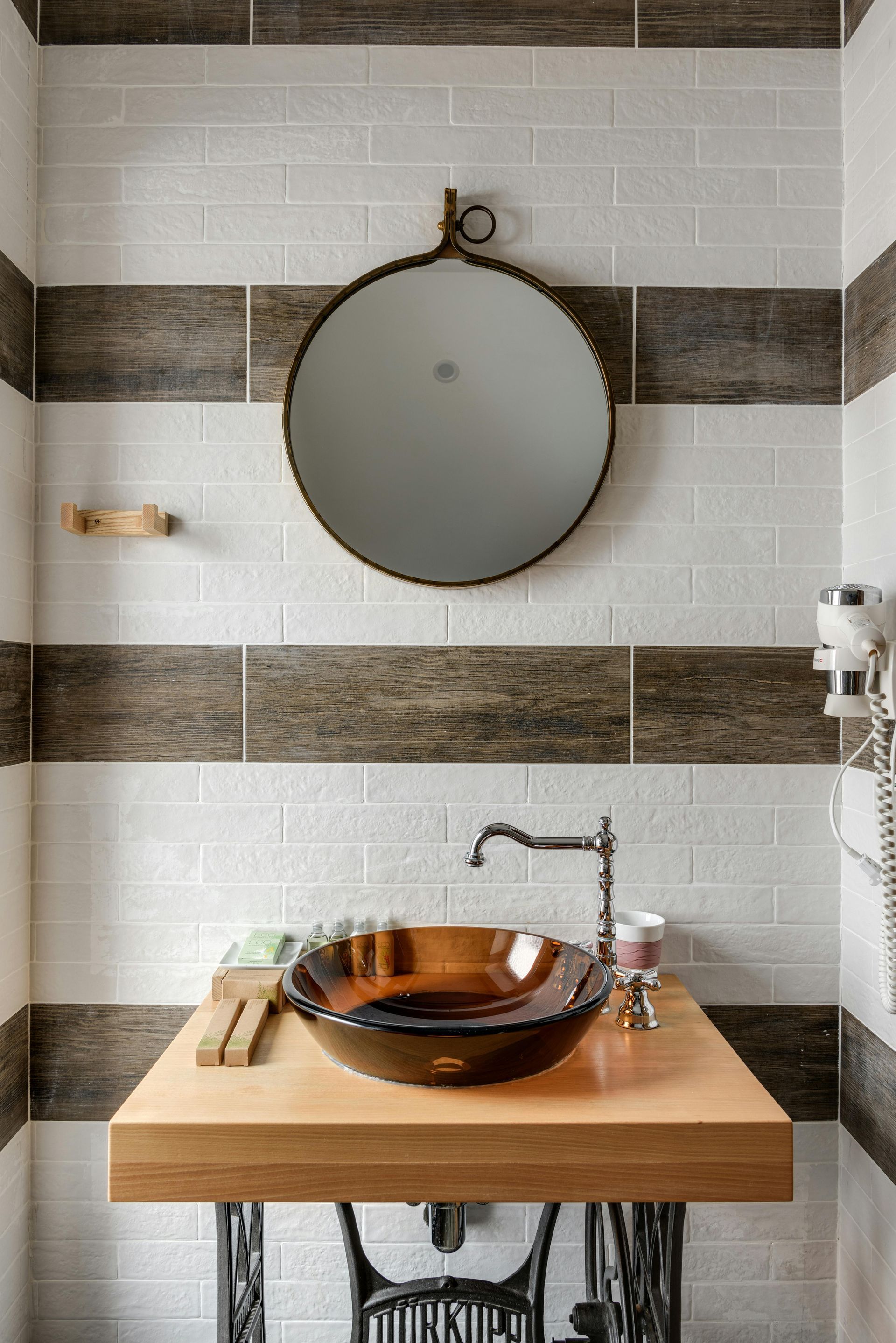Bathroom with a round mirror, brown vessel sink, and wood-topped vanity. Brown and white striped tile walls.