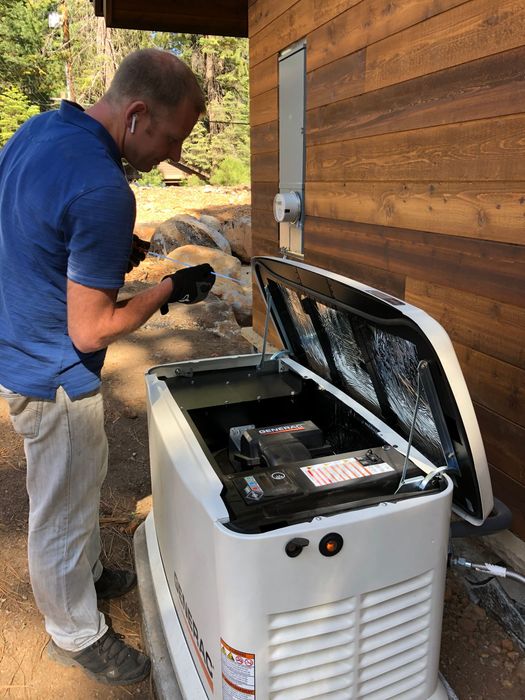 Man working on a white generator outside a building with brown siding. The generator's lid is open.