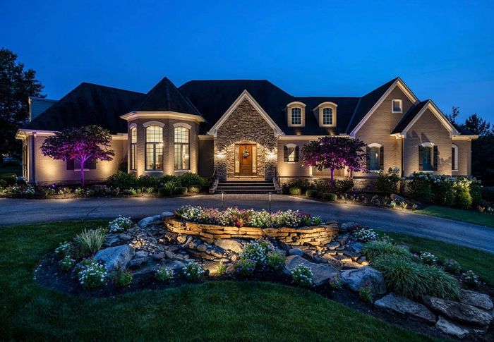 Elegant house at night, lit with exterior lights. Landscaping with illuminated water feature in front.