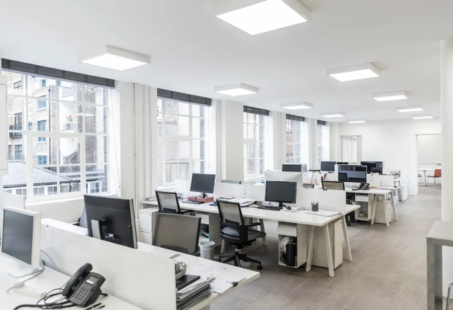 Bright, modern office with rows of white desks, computers, and windows overlooking a cityscape.
