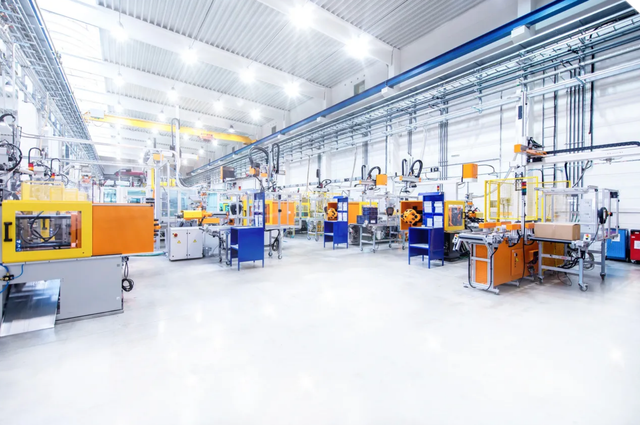 Factory interior with industrial machinery, orange and blue machines, white floor, bright overhead lighting.