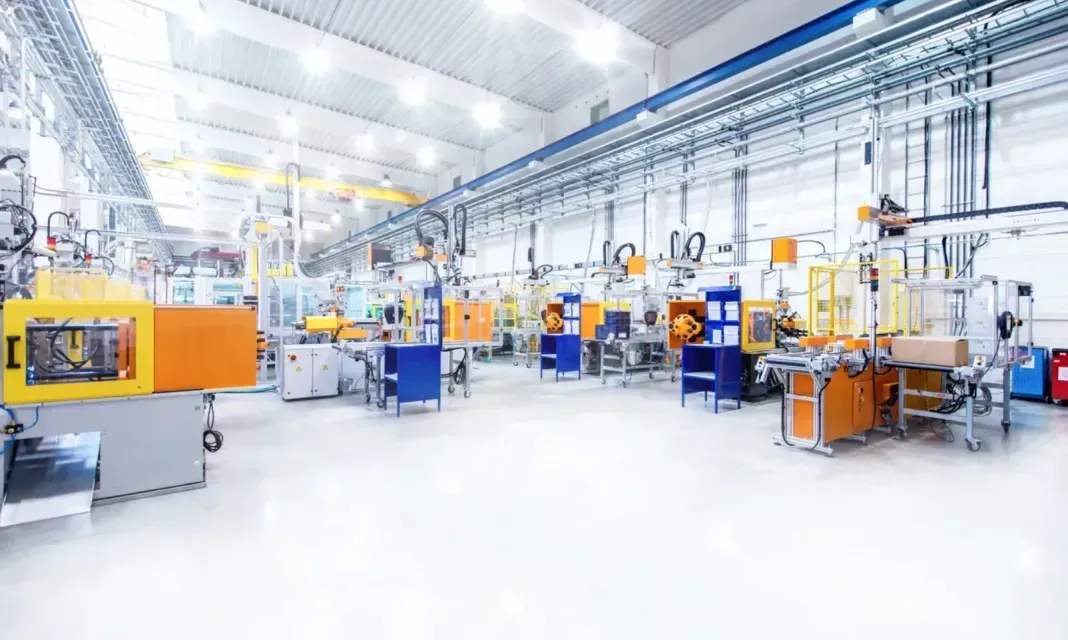 Factory interior with industrial machinery, orange and blue machines, white floor, bright overhead lighting.