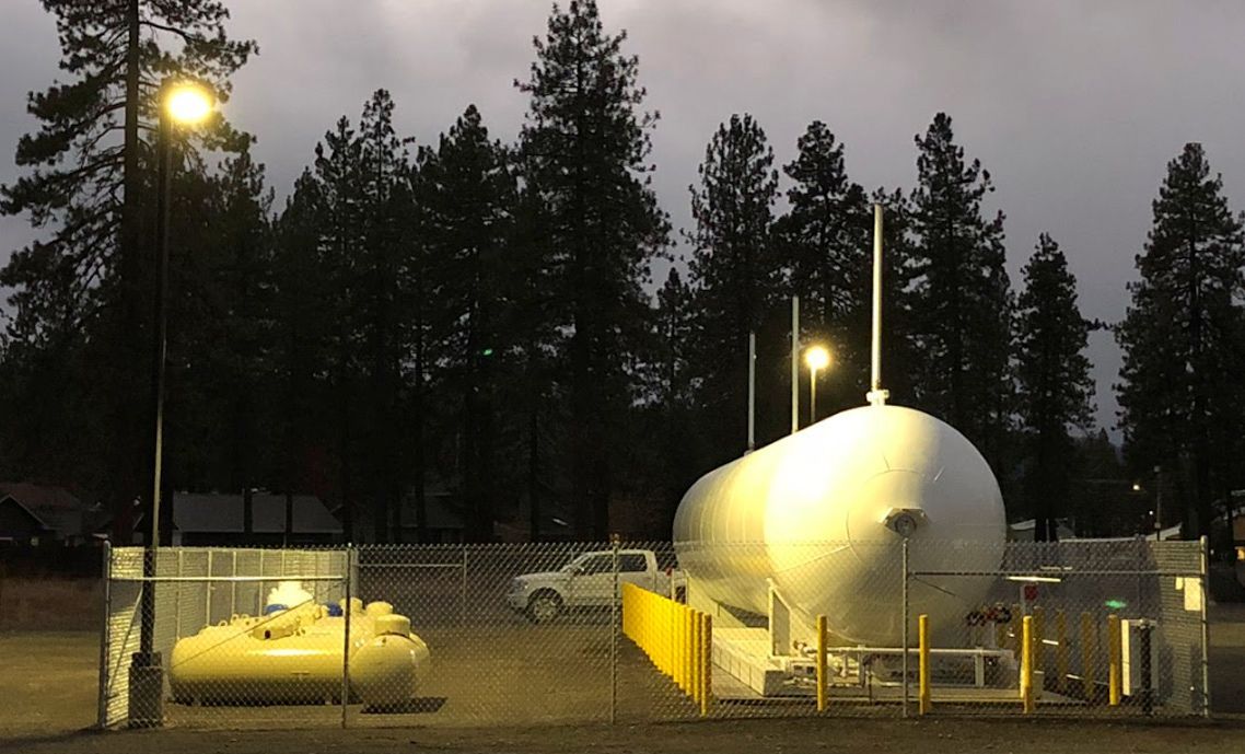 Large propane tank illuminated at dusk, enclosed by a fence, with a smaller tank and vehicle nearby.