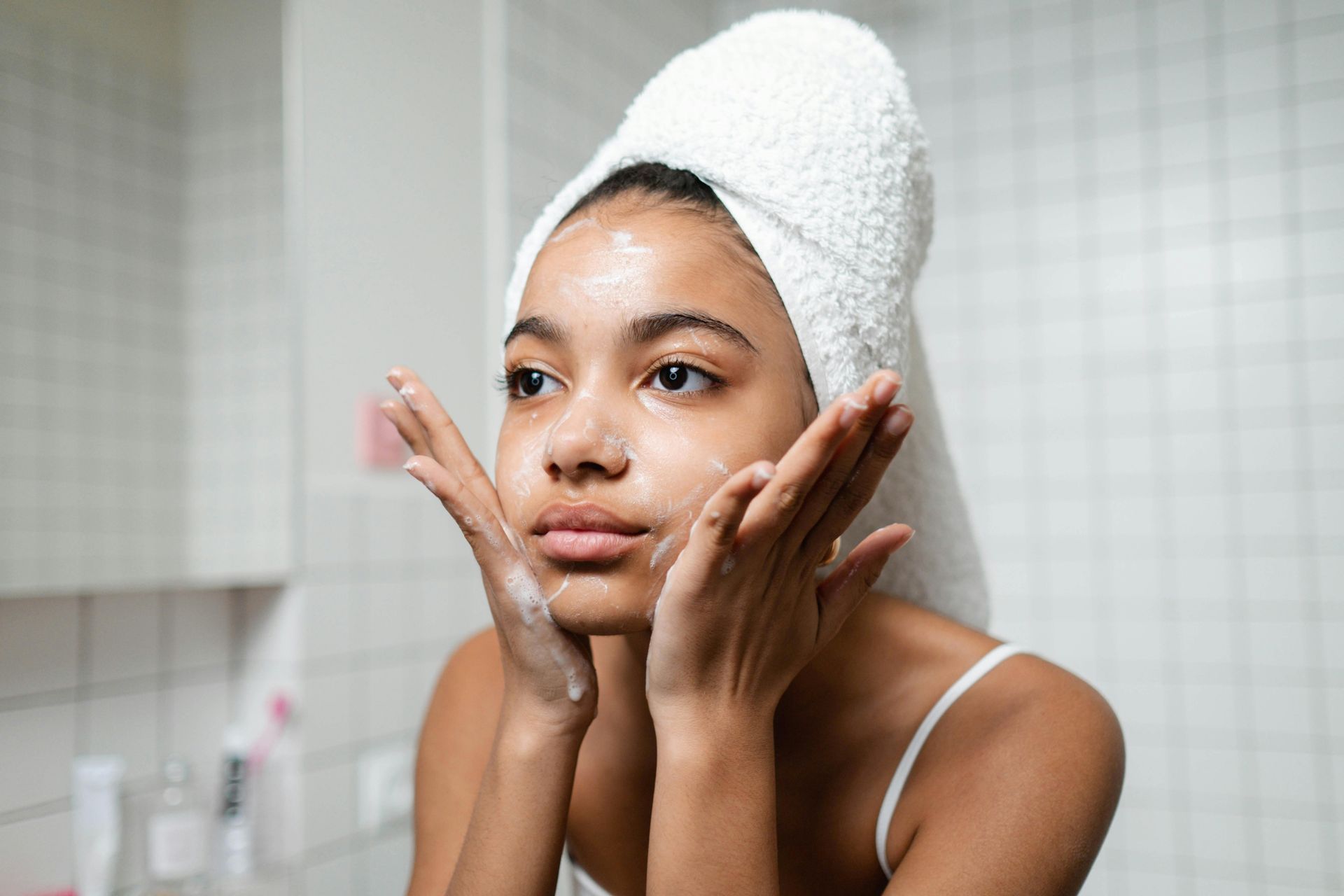 A woman with a towel wrapped around her head is washing her face in the bathroom.