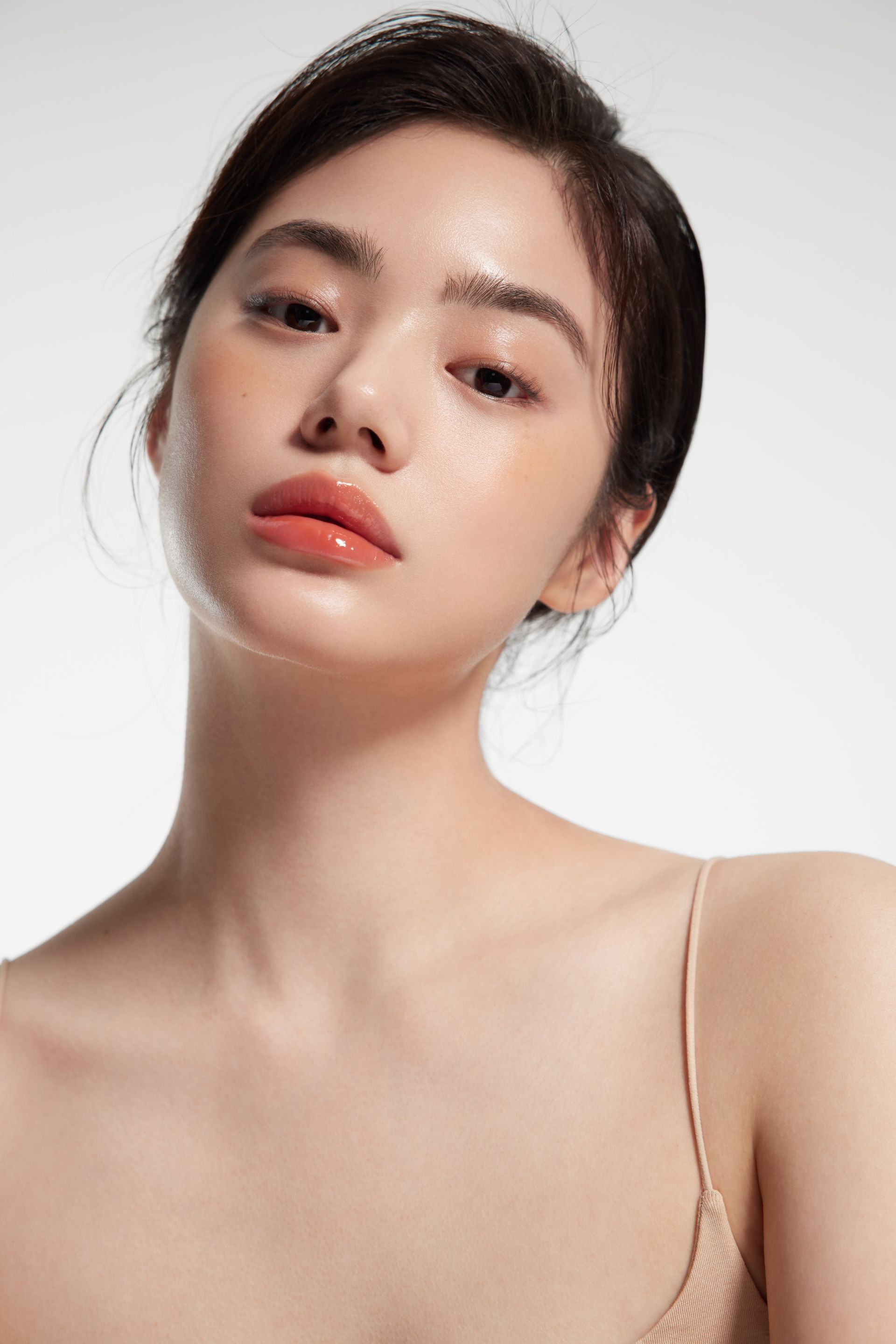 Woman with dark hair, minimal makeup, and a natural, glossy complexion, wearing a light spaghetti-strap top, looking towards the camera.