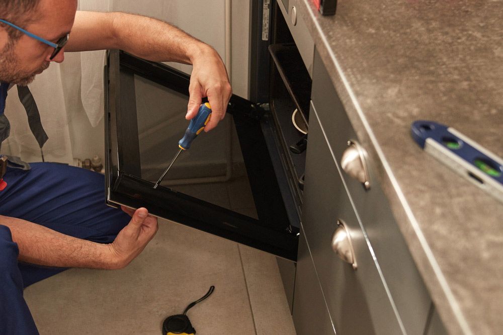 A Man is Fixing an Oven Door With a Screwdriver in a Kitchen — Patrick Lunney Home Appliance Service & Repairs in Maudsland, QLD