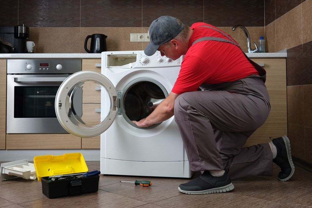 A Man is Fixing a Washing Machine in a Kitchen — Patrick Lunney Home Appliance Service & Repairs in Maudsland, QLD
