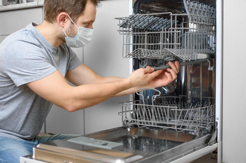 A Man Wearing a Mask is Fixing a Dishwasher — Patrick Lunney Home Appliance Service & Repairs in Maudsland, QLD
