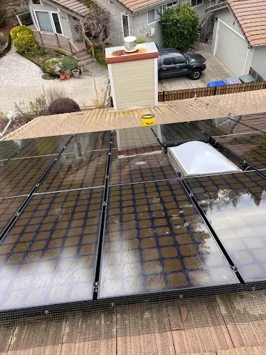Solar panels on a rooftop with a chimney, skylight, and partial view of a black car and surrounding houses.