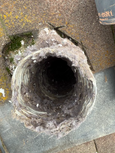 Exterior view of a clogged dryer vent exit covered in lint, against a weathered surface.