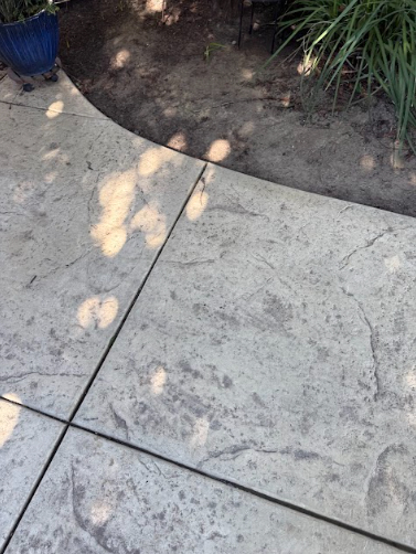 Textured gray concrete patio with dark lines, next to dirt and grass.