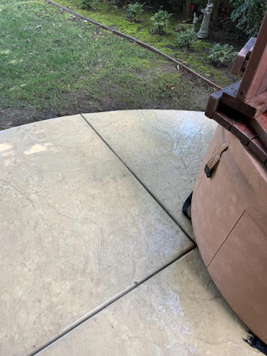 Concrete patio with a curved edge, adjacent to a grassy lawn. A wooden structure is partially visible.