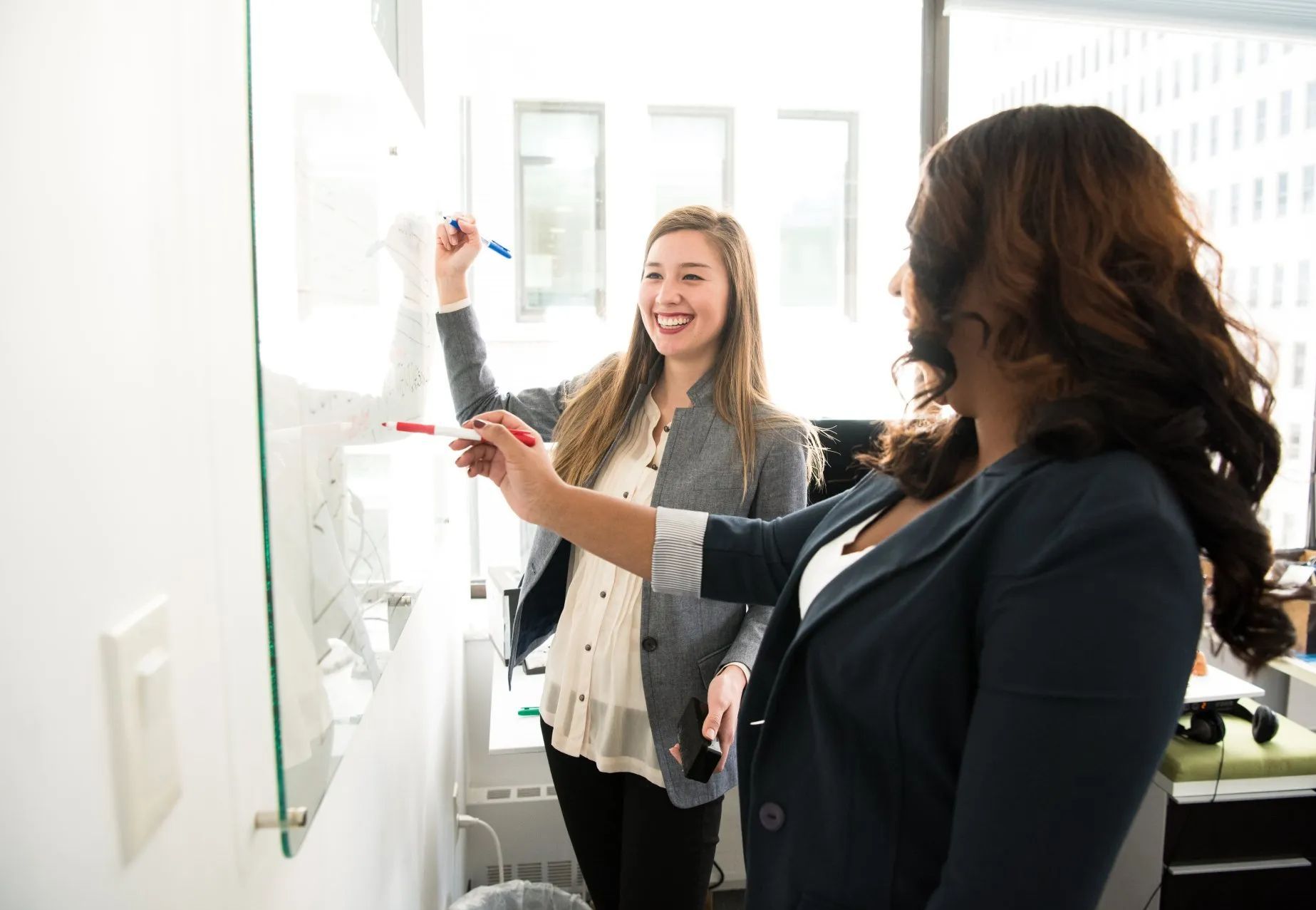 Two women are standing next to each other in front of a whiteboard.