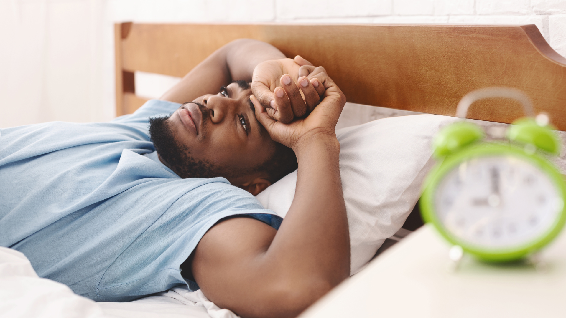 Man in bed, looking tired. Hand covering his eyes. Green alarm clock in the foreground.