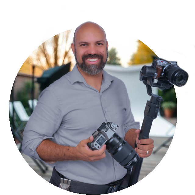 Bald man, gray shirt, holding two cameras, smiling, outdoor photo shoot.