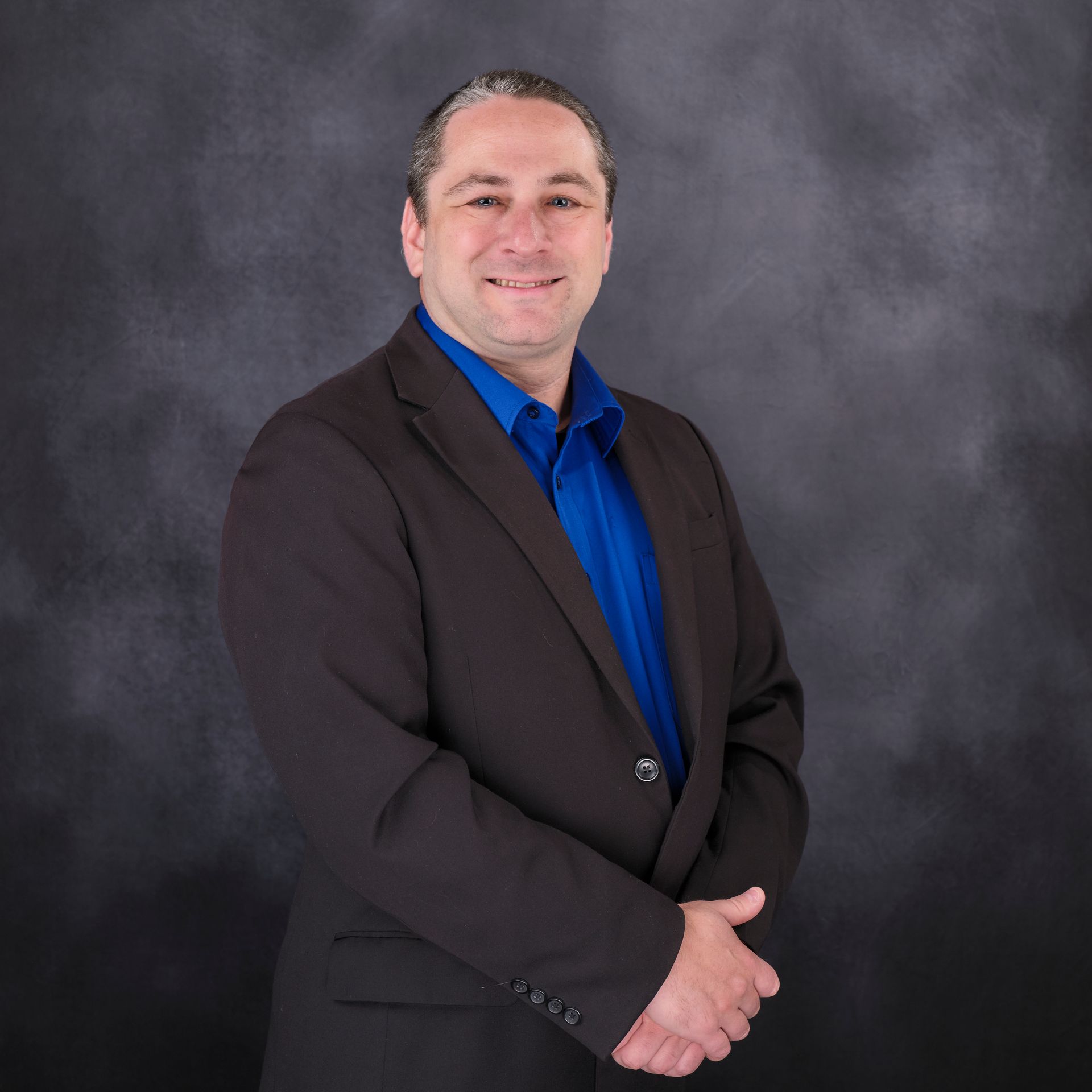Man in a suit smiling, wearing a blue shirt. Posing in front of a textured, dark background.
