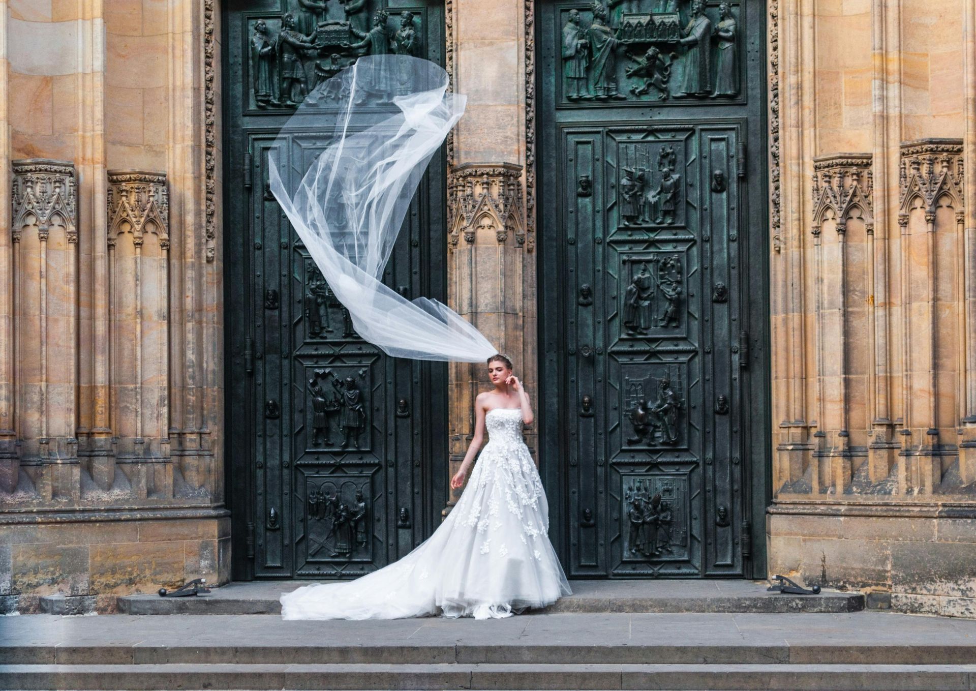 A bride in a wedding dress is standing in front of a door with her veil blowing in the wind.