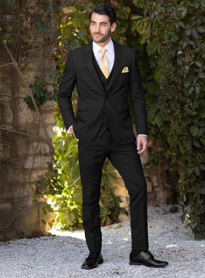 A man in a black suit and yellow tie is standing in front of a stone wall.