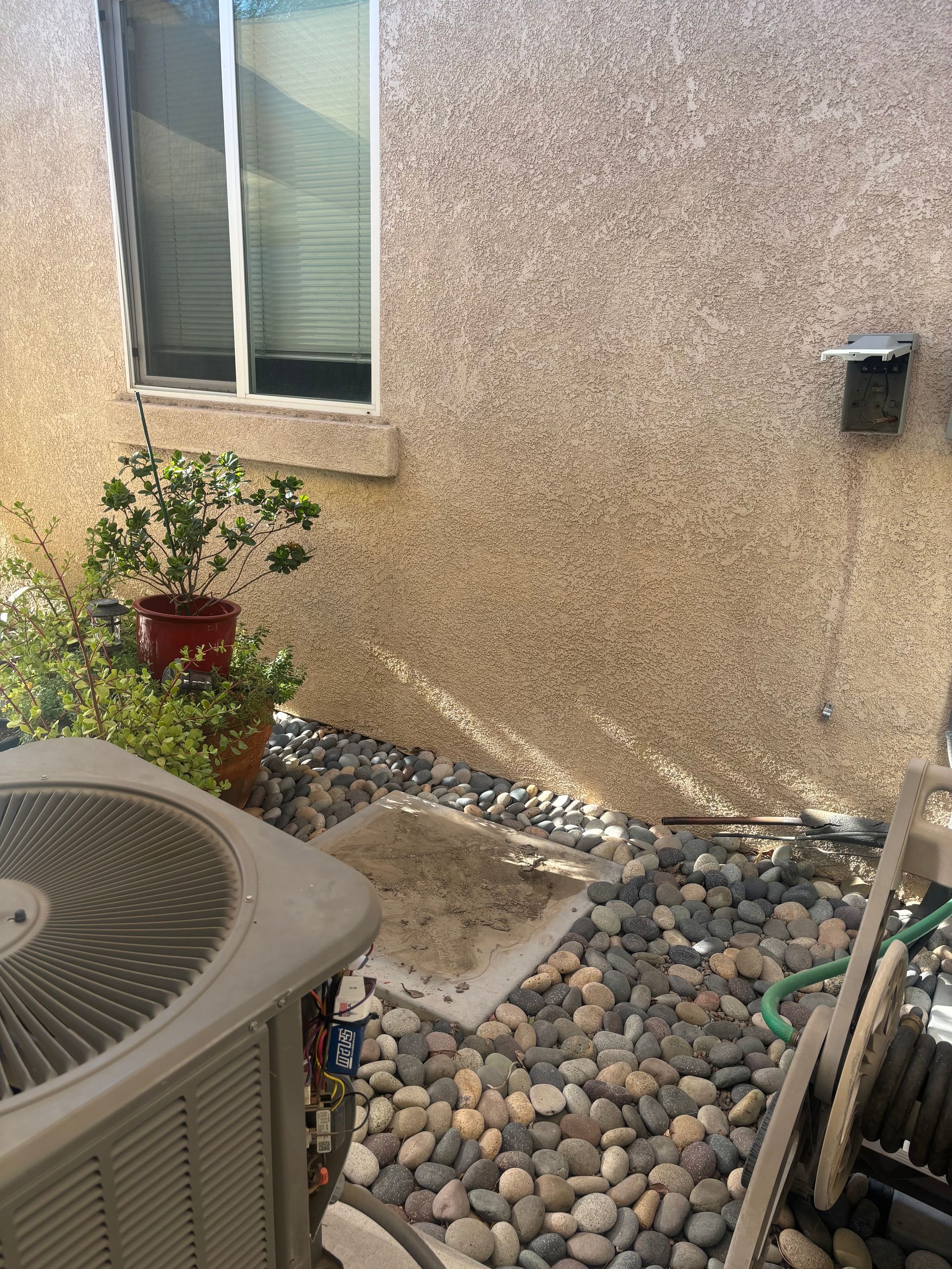 Exterior view of a beige stucco wall with a window, plants, and an air conditioning unit surrounded by pebbles. A chair is visible to the right.