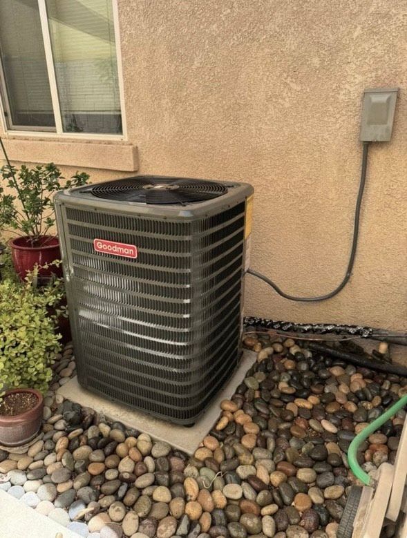An outdoor Goodman air conditioning unit next to a beige wall, sitting on a concrete pad surrounded by pebbles.