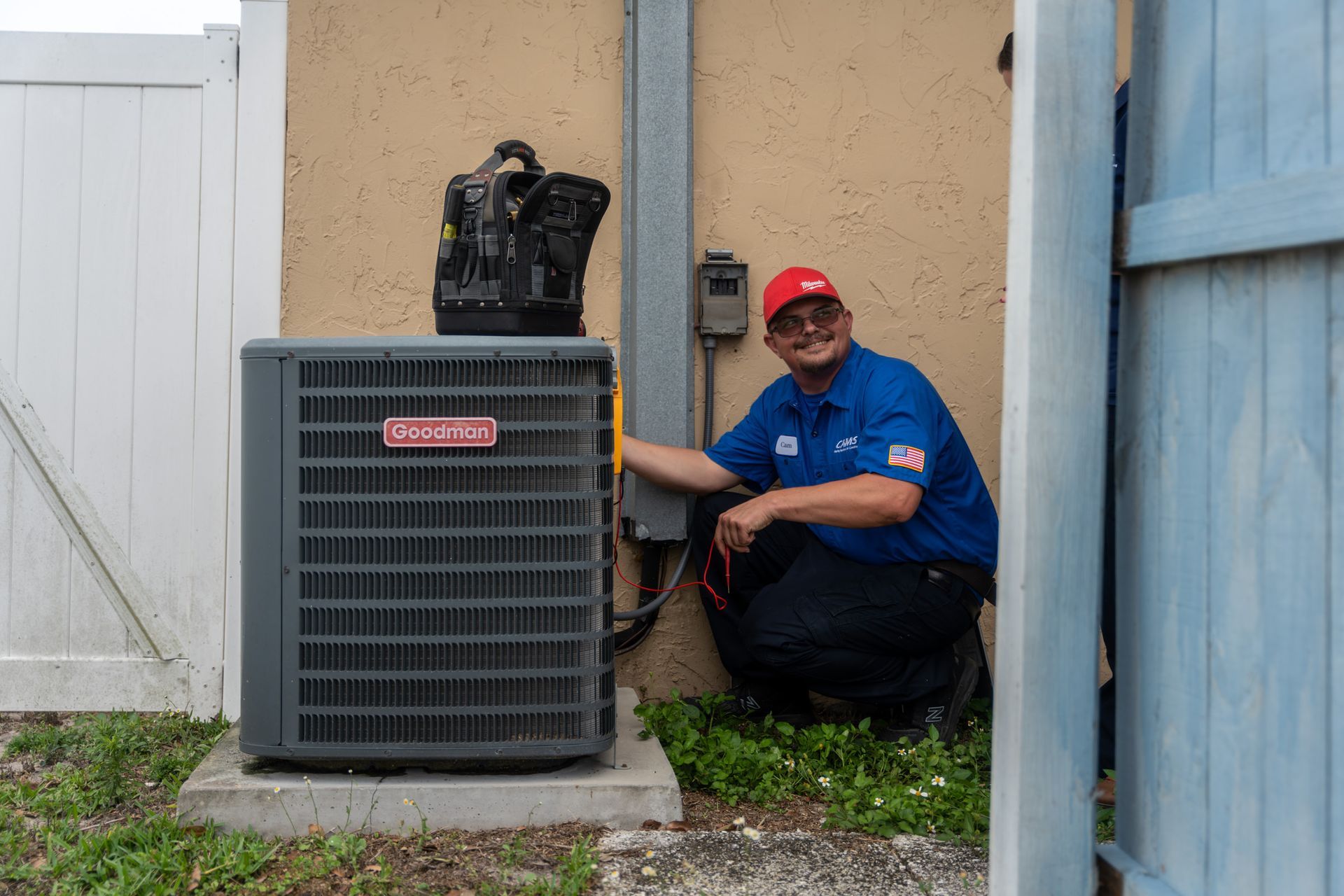 HVAC technician next to an air conditioning unit. He's kneeling, smiling, and wearing a red hat and blue shirt.