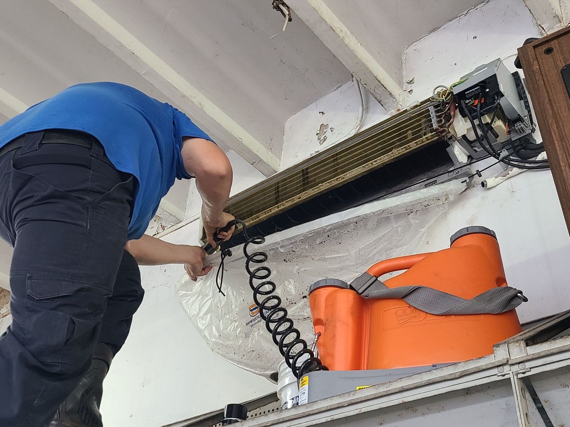 Person in blue shirt repairing an air conditioning unit; orange container below.