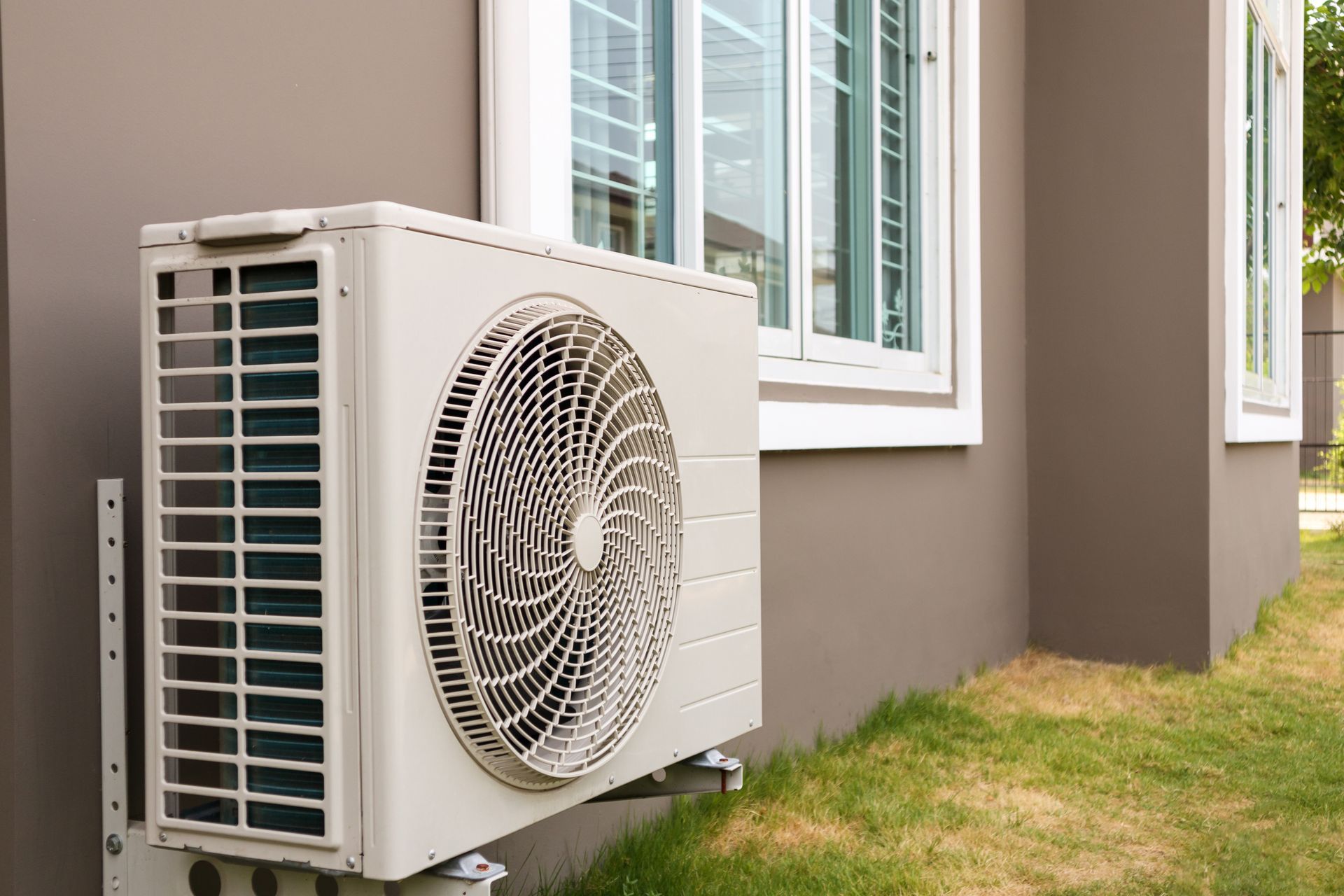 A white air conditioner is mounted on the side of a house next to a window.