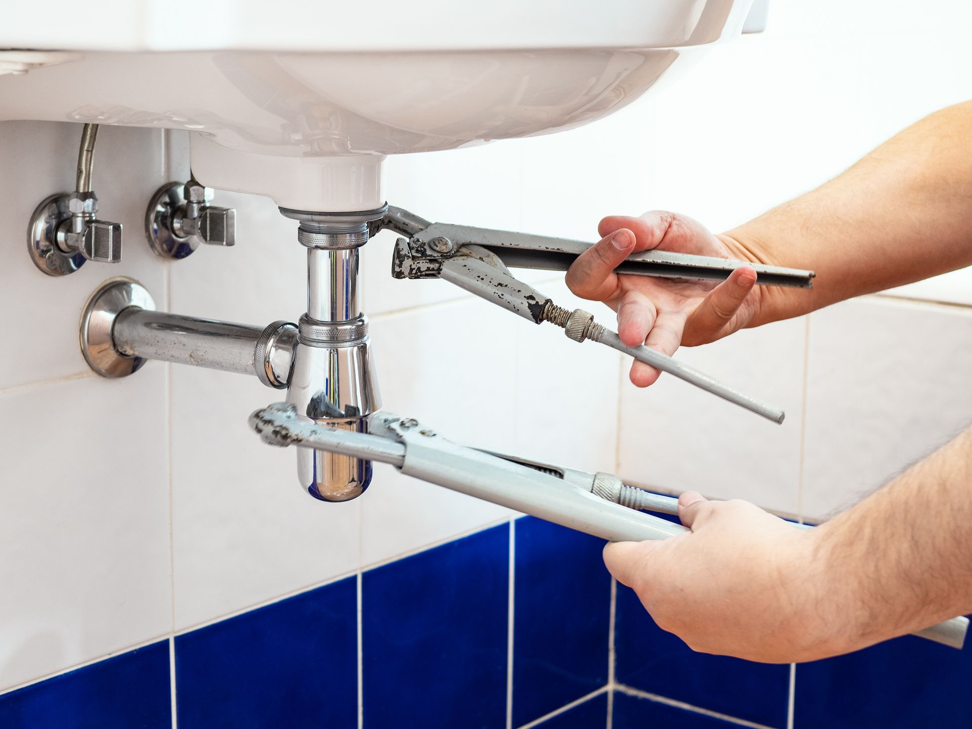 Person using two wrenches to work on plumbing under a white sink in a bathroom with blue and white tiles.