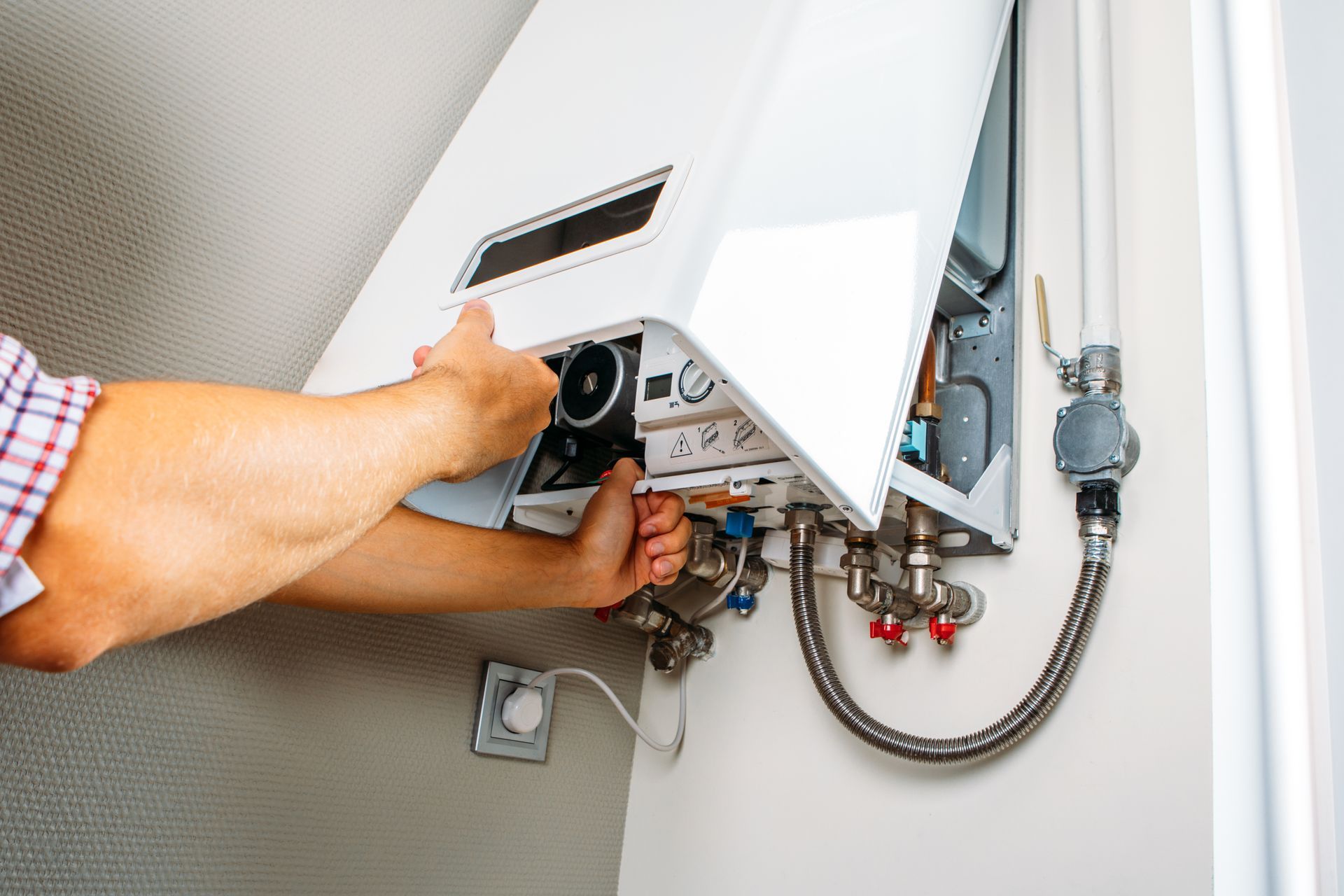 Person's hands working on a white wall-mounted boiler, near pipes and a wall outlet.