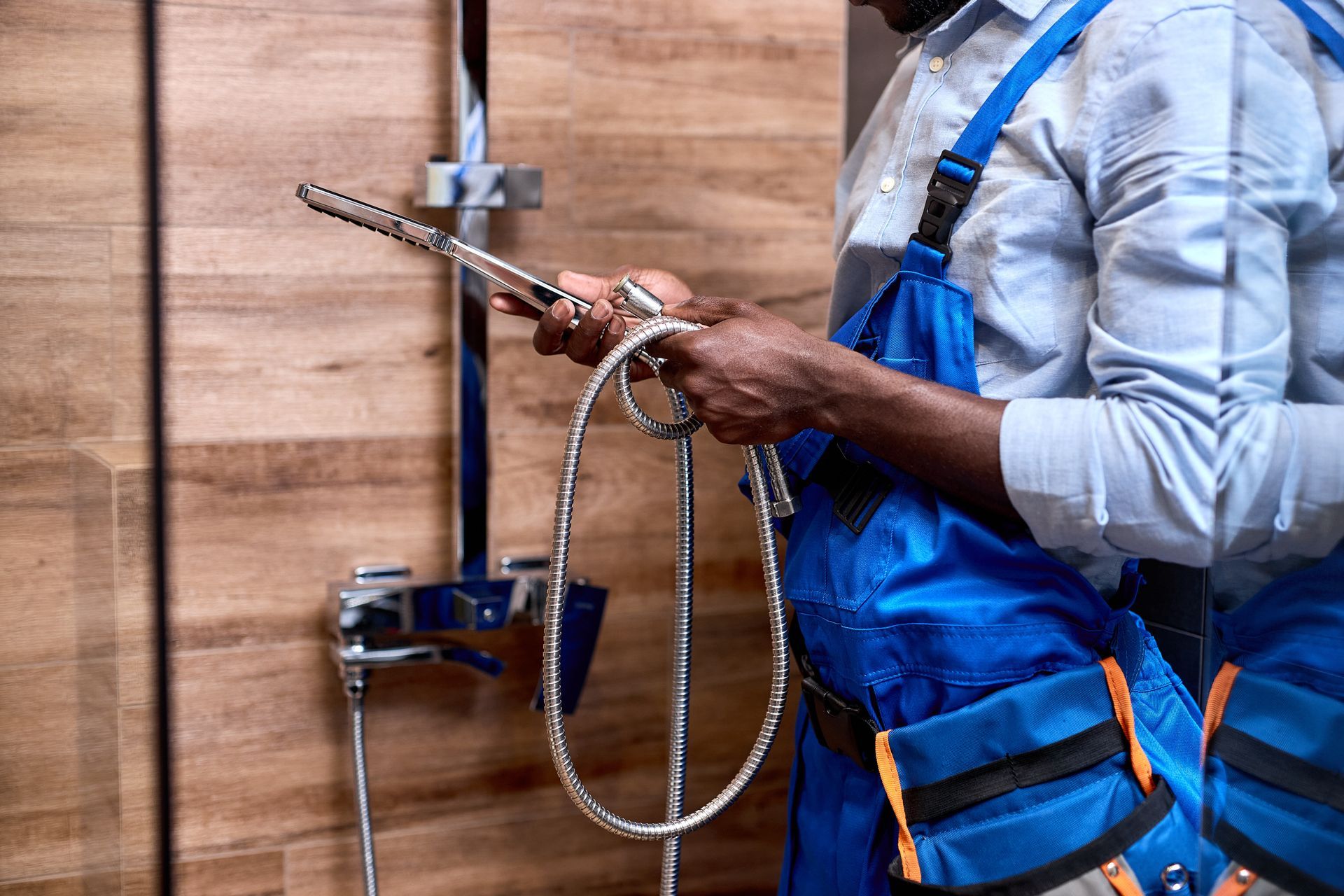 Plumber examining shower hose, standing near a wooden-paneled shower wall, wearing blue overalls and a tool bag.