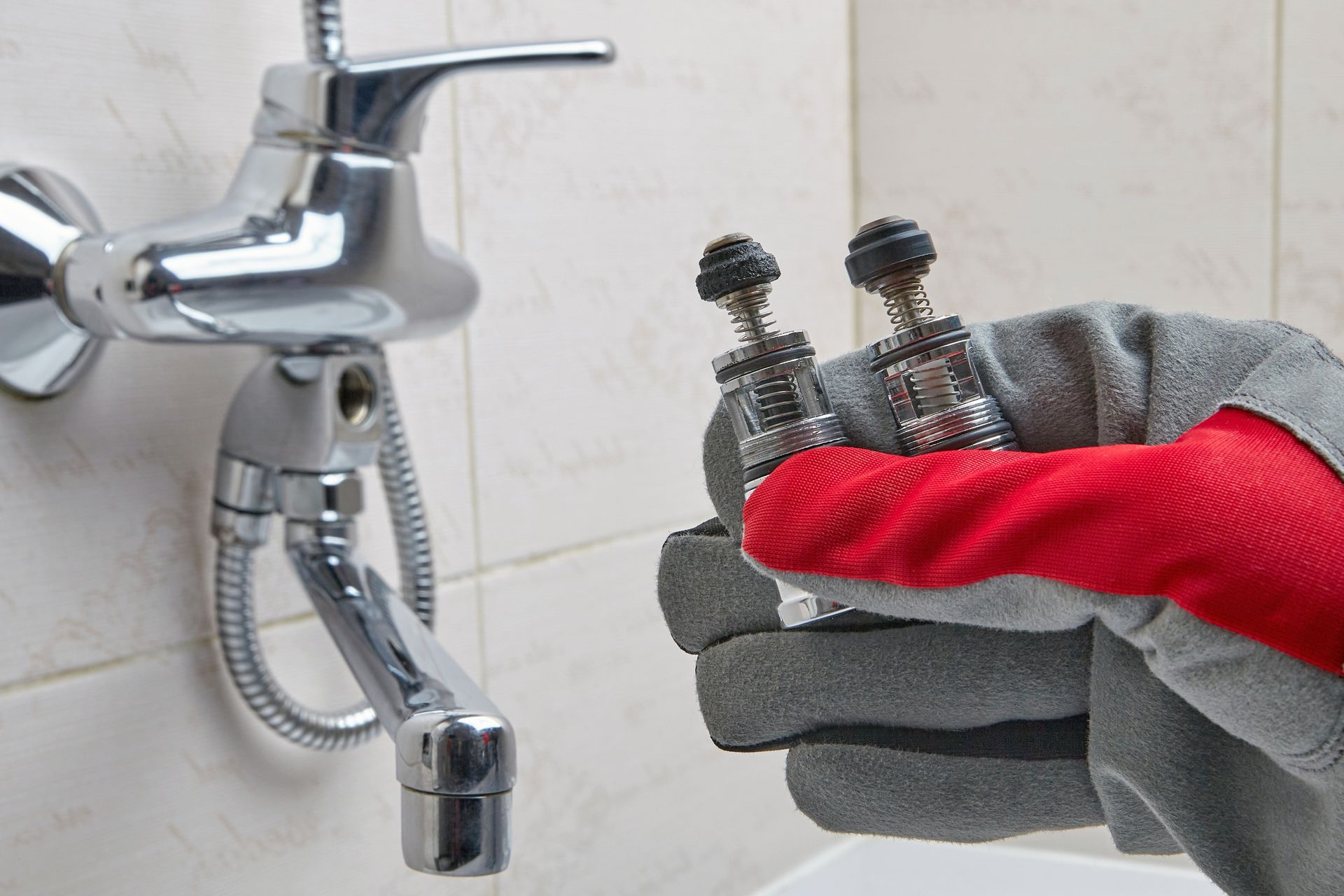 Gloved hand holding two removed faucet valve stems, near a chrome shower faucet on a tiled wall.