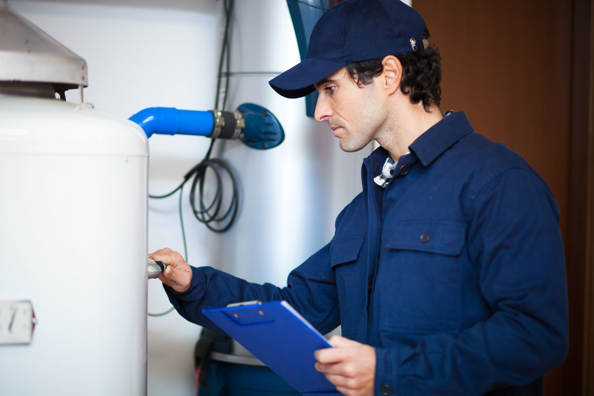 Plumber in blue uniform examines a water heater, holding clipboard.