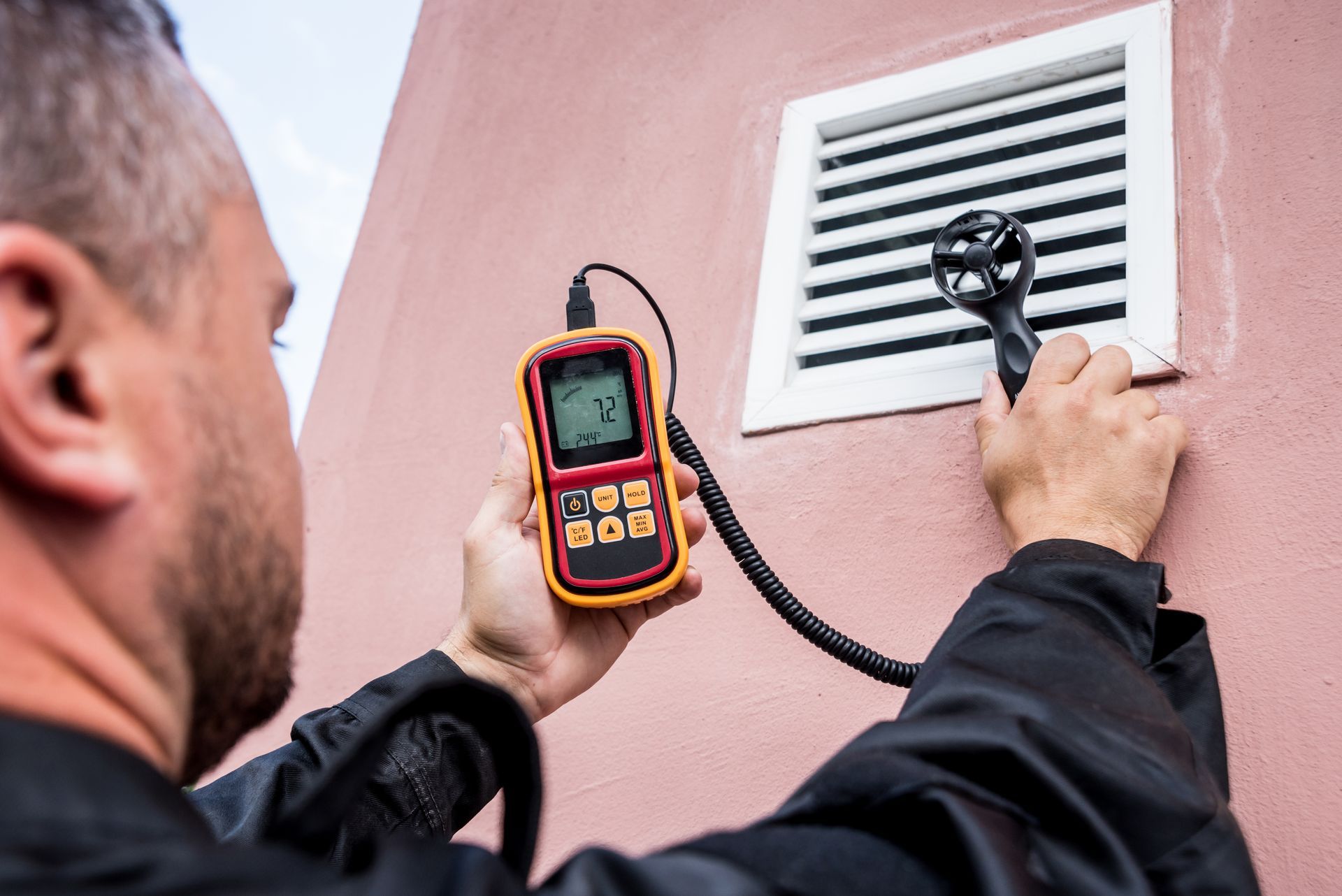 Man using a handheld anemometer to measure airflow from a vent on a pink wall.