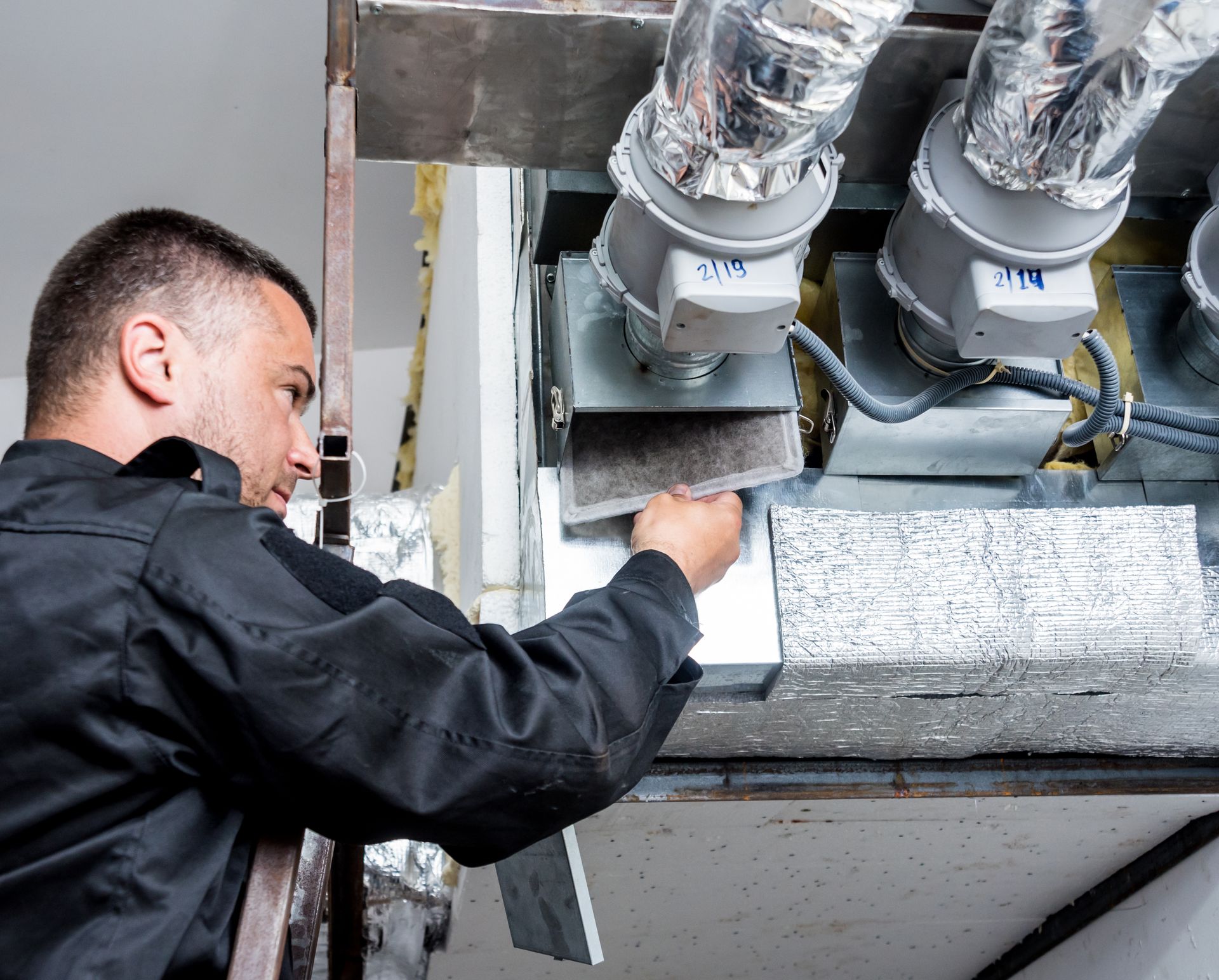 HVAC technician, in black uniform, inspecting an air duct system.