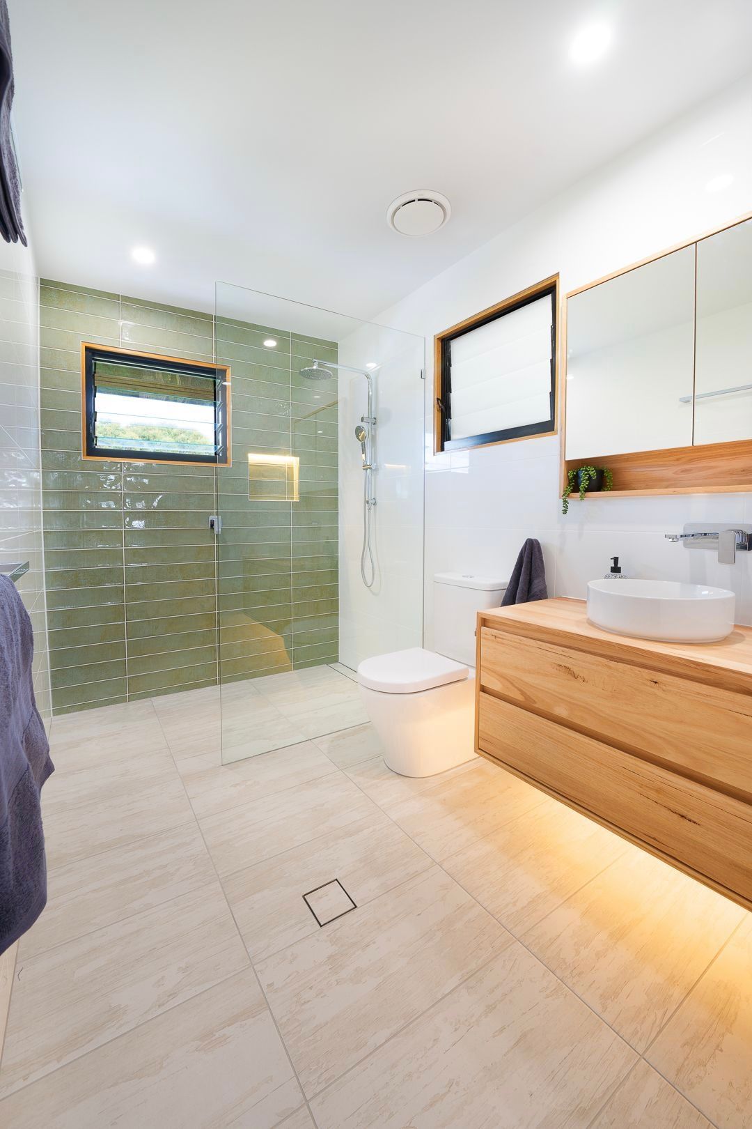 A modern bathroom with a light wood vanity, a vessel sink, a white toilet, and a glass-enclosed shower with green tiles.
