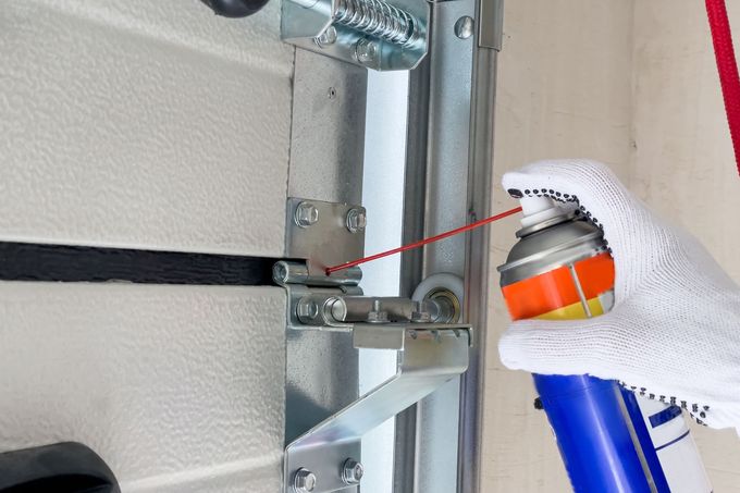 Gloved hand sprays lubricant onto a garage door hinge and metal track assembly