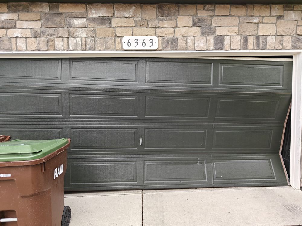 Dark green garage door with house number 6363, stone wall above, and a trash bin at left.