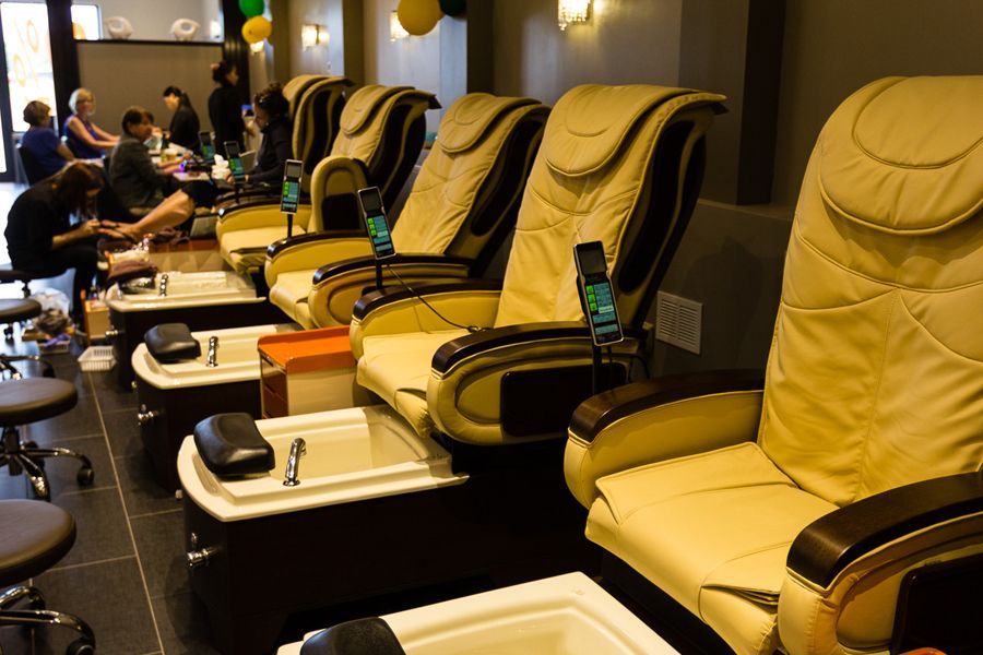 Nail salon interior: beige massage chairs, foot baths, dark floor, customers getting manicures and pedicures.