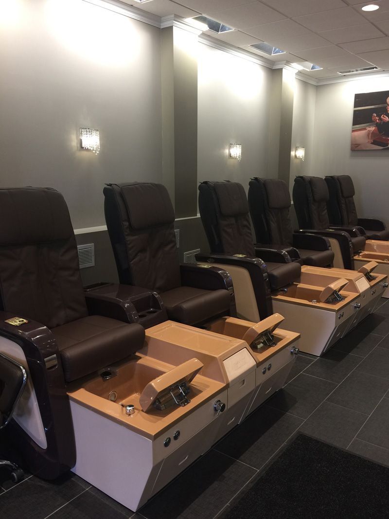 Four brown pedicure chairs in a nail salon, each with a foot bath. Gray walls, tiled floor.