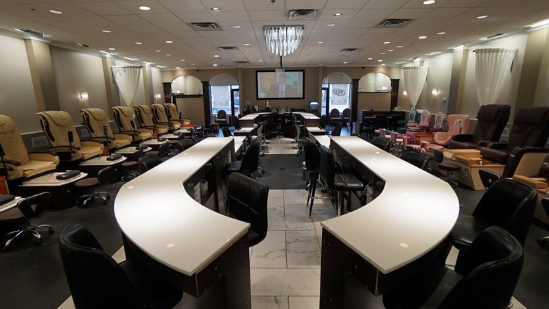Nail salon interior with U-shaped counter, manicure stations, and pedicure chairs. Neutral tones, bright overhead lighting.